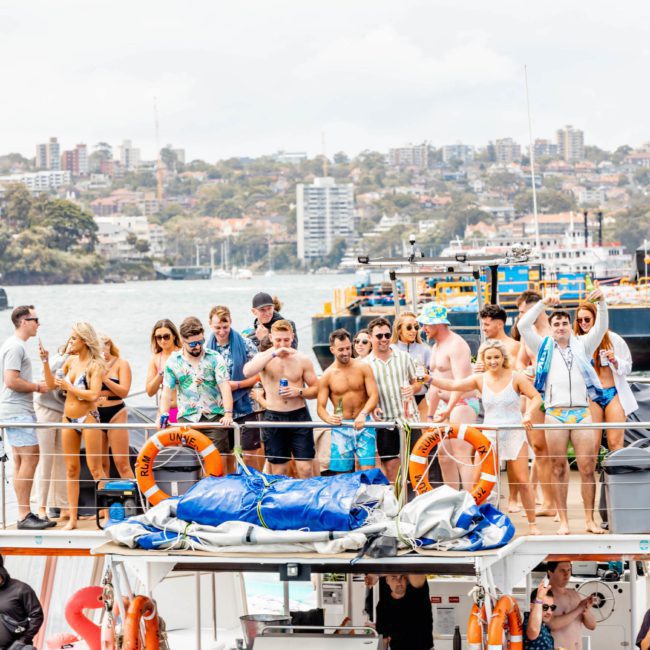 A group of people in swimsuits and summer clothing stand on the deck of a boat, with a waterfront cityscape in the background, enjoying a vibrant atmosphere typical of catamaran parties in Sydney.