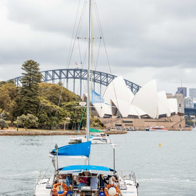 A catamaran sails in Sydney Harbour with the Sydney Opera House and Sydney Harbour Bridge in the background on a cloudy day, perfect for corporate boat events or a private yacht charter.