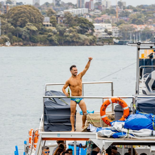 A man in swim trunks stands on the upper deck of a boat, gesturing, with a cityscape visible in the background. Several people are gathered on the lower deck, enjoying a corporate boat event in Sydney Harbour.
