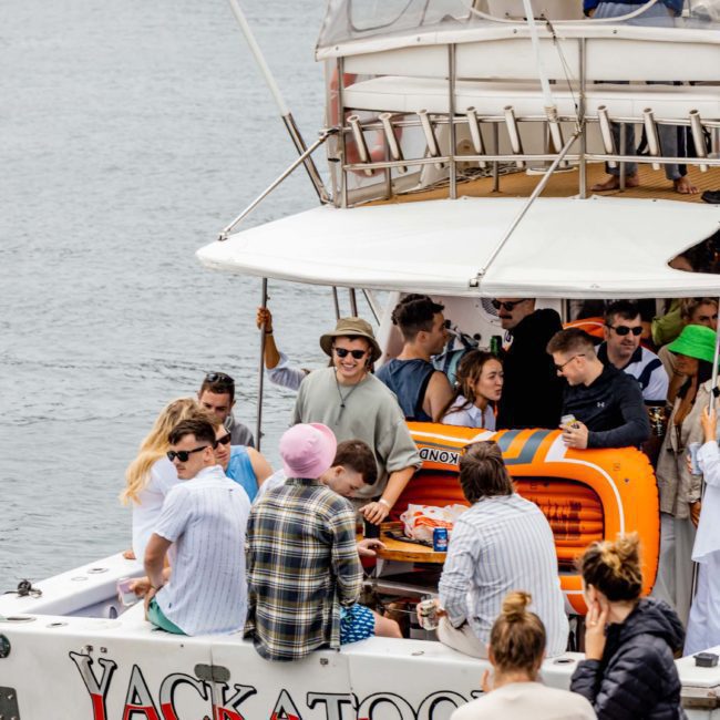 A group of people socialize on a catamaran named "Jackatoo" anchored on the water, with a large orange cooler and various items on board. Perfect for corporate boat events in Sydney, this setup promises an unforgettable experience.
