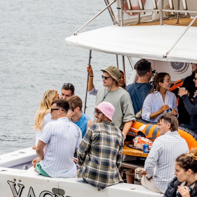 A group of people socialize on a boat named "Yackatoon" floating on the water. Some are standing and talking, while others are seated. The backdrop features calm water, making it perfect for corporate boat events Sydney or even considering DJ boat hire Sydney for added entertainment.