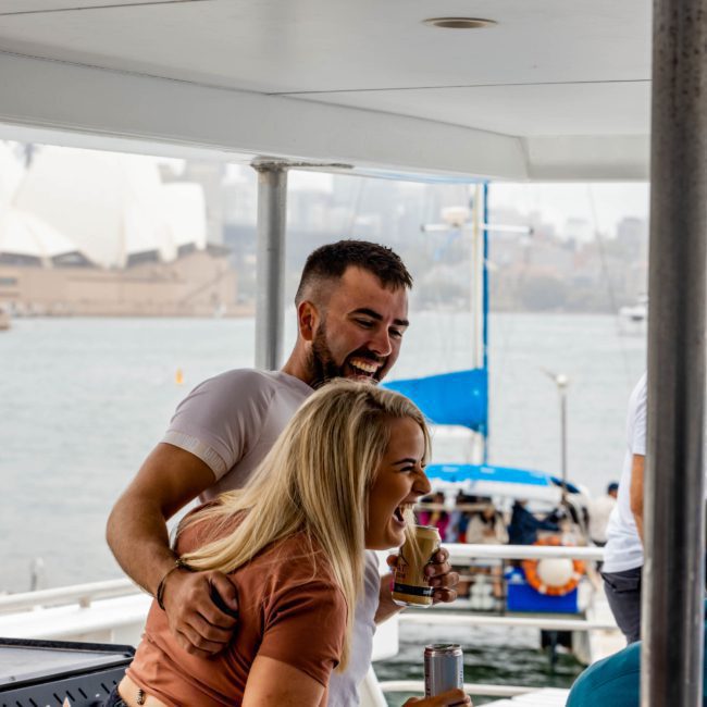 A man and a woman laugh and hold drinks on a boat with a cityscape and the Sydney Opera House gleaming in the background, enjoying their luxury yacht hire in Sydney.