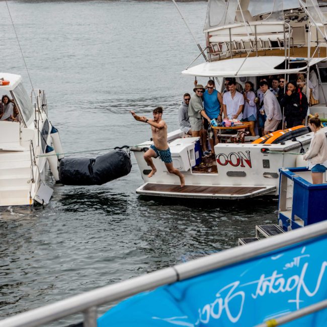 A man in swim shorts jumps from a boat towards another vessel as several people watch from the deck. The lively scene, part of a Sydney boat party hire, takes place on a body of water with trees visible in the background.