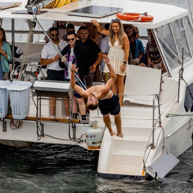 A man dives off the side of a luxury yacht into the sparkling waters while several people on the deck watch and smile, experiencing the thrill of a private yacht charter in Sydney Harbour.