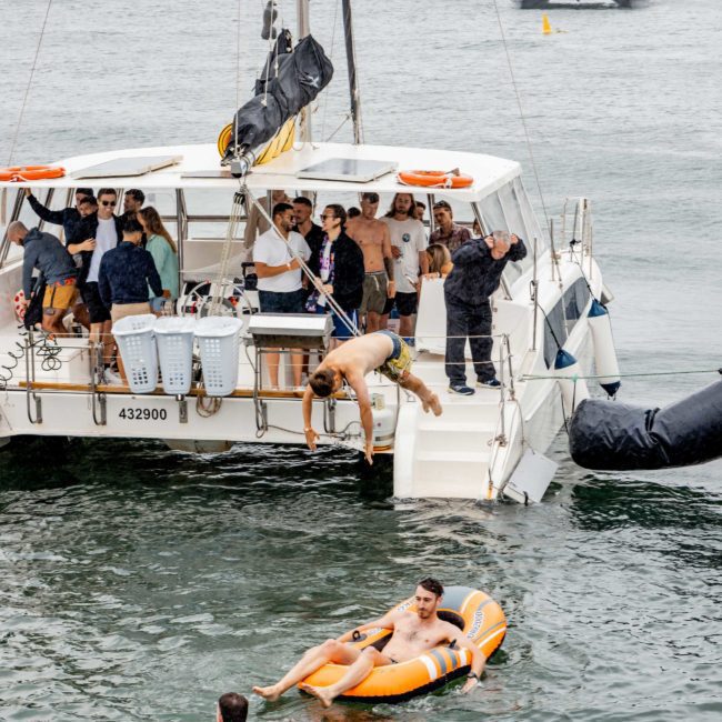 A group of people are enjoying a private yacht charter on Sydney Harbour, with some playing near the water. One person is about to jump in, and two individuals are floating on inflatables nearby. Another boat is visible in the background.