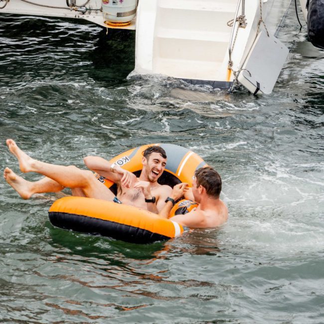 Two men float on an inflatable ring in the water near the back of a boat, laughing and splashing. Additional people and boat parts are visible in the background, suggesting a fun Sydney boat party hire.