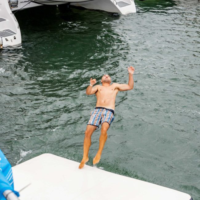 A person in swim shorts falling backward from a boat onto a floating mat on the water, with onlookers watching from surrounding boats during a Sydney boat party hire.
