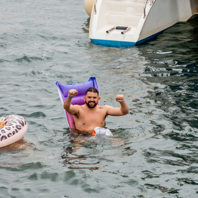 A man in the water holds his arms up in a celebratory gesture while sitting on a purple float near a luxury yacht hire Sydney. Another circular float is partially visible.