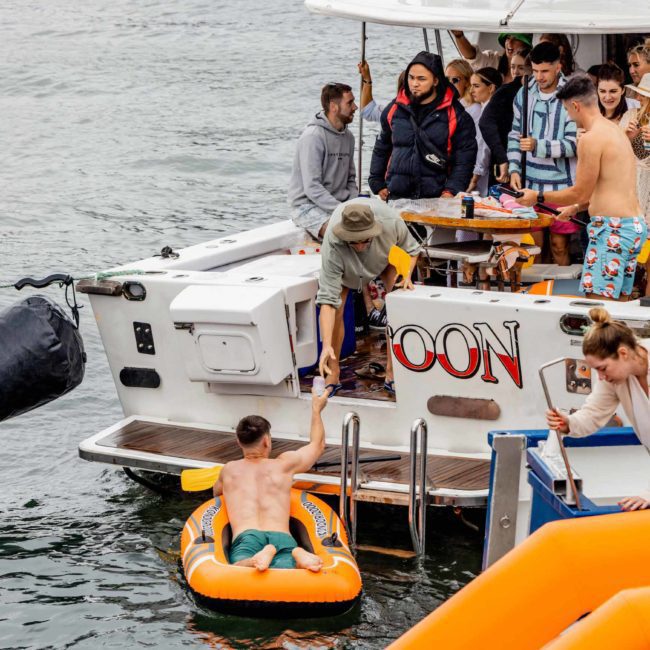 People enjoying a Catamaran party Sydney, some on the boat and others on an inflatable raft or in the water. The boat has "OON" written on the back.