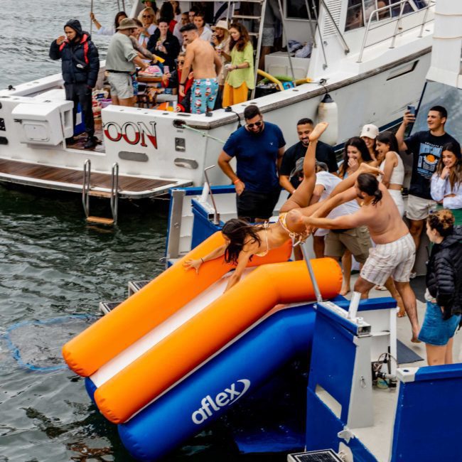 Guests are enjoying a Catamaran party in Sydney, with two individuals sliding down an inflatable slide into the water. Other guests are standing and watching from two moored boats.
