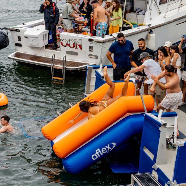 People enjoy a corporate boat event in Sydney, with some sliding off inflatable water slides and others swimming or socializing on deck.