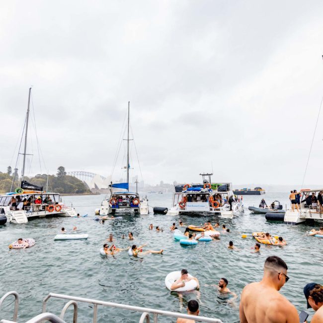 People are swimming and floating on inflatables in the water, surrounded by several boats. The scene is lively, with individuals enjoying a day out on the water, perhaps as part of a Sydney boat party hire.