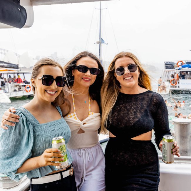 Three people are standing on a boat, wearing sunglasses and holding canned drinks. Other boats and people in the water are visible in the background, creating the perfect vibe for a Sydney boat party hire or a private yacht charter on Sydney Harbour.