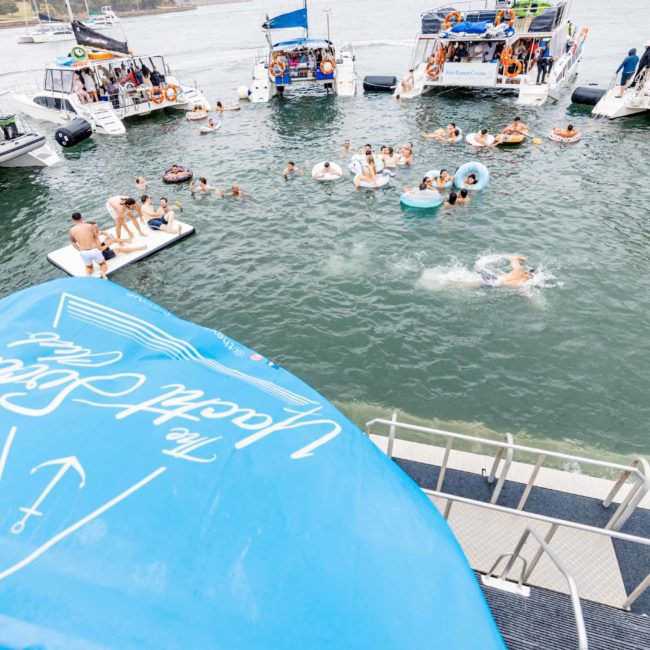People are swimming and lounging on floaties in the water, surrounded by several luxury yachts docked nearby, creating the perfect atmosphere for a Sydney boat party hire.