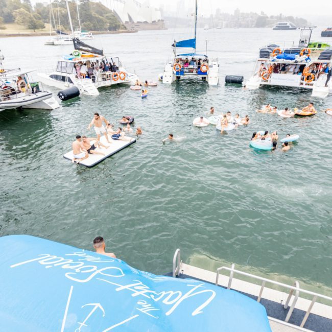 People enjoying a sunny day on boats in a harbor, swimming, and floating on inflatables in the water. Multiple boats are anchored close together surrounded by lush greenery. Perfect for a Sydney boat party hire or corporate boat events Sydney.
