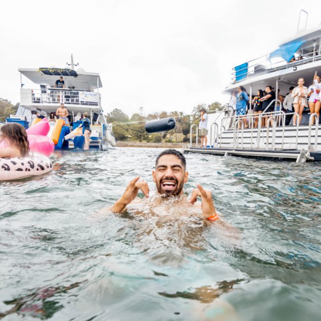 A man is swimming and smiling in the water, with two boats and several people visible in the background, suggesting a lively Catamaran party Sydney atmosphere.