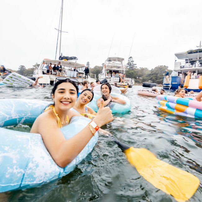 A group of people in inflatable tubes are enjoying a day on the water, surrounded by various boats. One person in the foreground is giving a thumbs up while paddling with an oar. It's the perfect scene for a Sydney boat party hire or even considering a private yacht charter on Sydney Harbour.