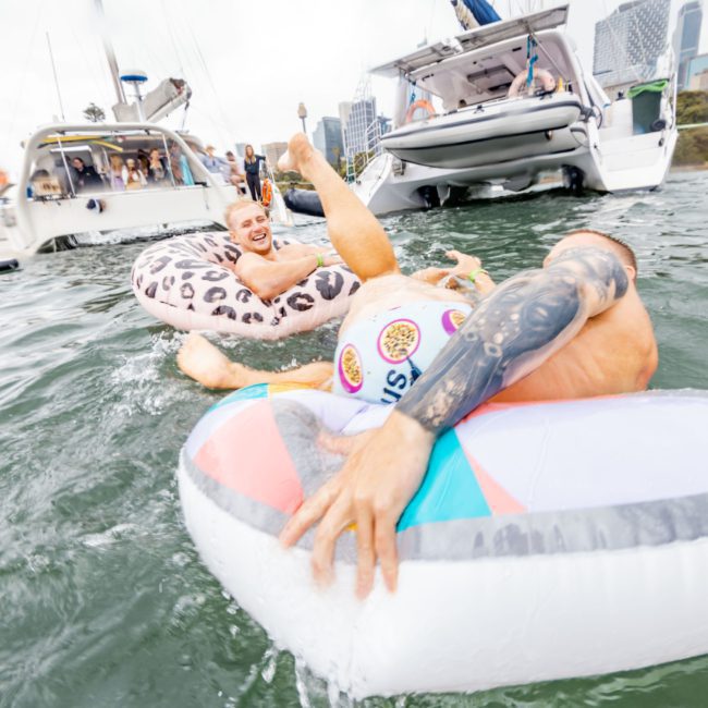 Two people in inflatable rings enjoy the water near anchored boats, with city buildings visible in the background. One person is partially submerged, and the other is laughing, capturing the joy of a Sydney boat party hire.
