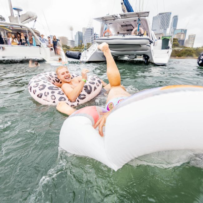 A person is lying on an inflatable float in a body of water, giving a thumbs-up. Boats and other people are seen in the background. Consider adding to the fun with a private yacht charter on Sydney Harbour for an unforgettable experience.
