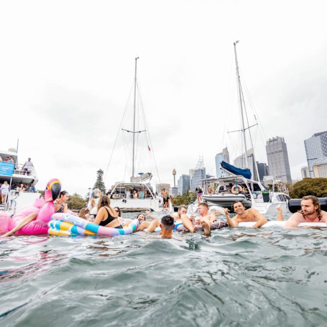 A group of people relax on inflatable floats in the water near several docked boats, with a city skyline in the background, enjoying a luxury yacht hire Sydney experience.