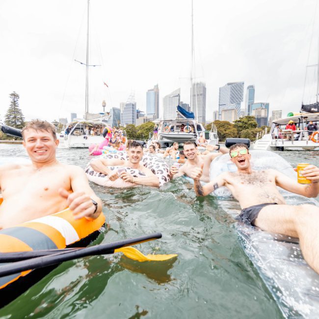 Group of young men enjoying a day on the water near boats and city skyscrapers, floating on inflatables and smiling at the camera. They’re making the most of their Sydney boat party hire, with vibrant scenes of fun against the backdrop of stunning views.