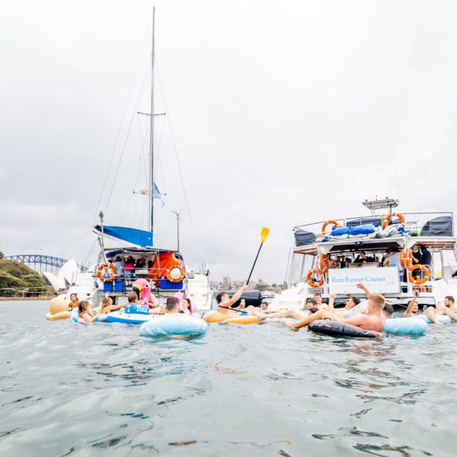 People floating on inflatable tubes in the water near anchored sailboats and a ferry on a cloudy day, enjoying a luxury yacht hire Sydney experience.