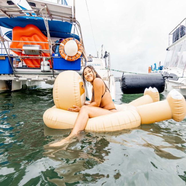 A woman in a swimsuit relaxes on an inflatable raft shaped like a face in the water, with private yacht charters cruising nearby.