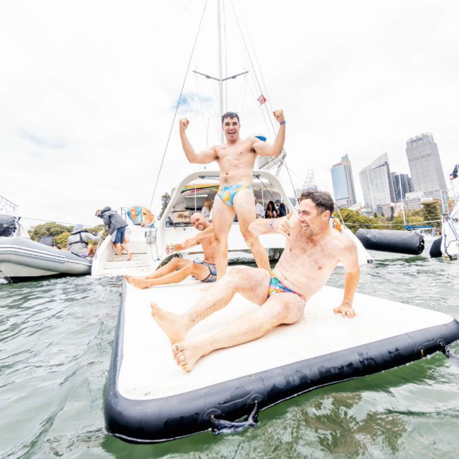 Three men in swim trunks are enjoying a day on the water, with two on an inflatable pad and one standing, flexing his muscles. Two boats, perhaps part of a luxury yacht hire Sydney experience, are in the background near a city skyline.