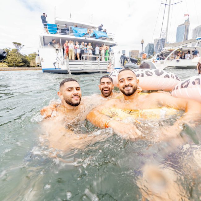 Three men in the water smile at the camera with a boat and other people in the background. The water is calm and the scene exudes joy and liveliness, perfect for a Sydney boat party hire.