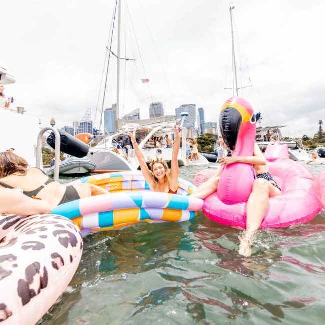 People on inflatable floats having fun in the water near boats, under partly cloudy skies with city buildings in the background. Consider a luxury yacht hire Sydney experience to elevate your day on the water.
