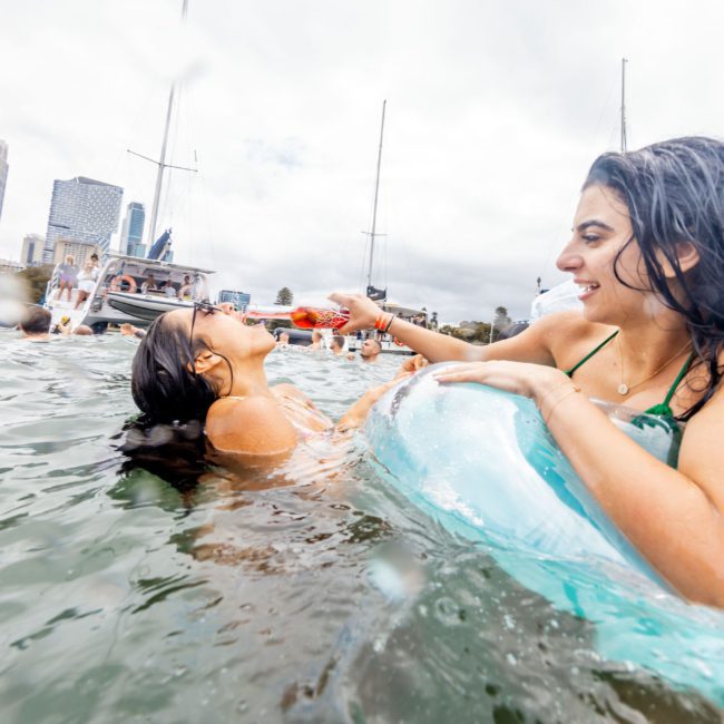Two women enjoy time in the water, one in an inflatable ring and the other swimming while being given a drink. Boats and a city skyline are visible in the background, hinting at a luxurious yacht hire Sydney experience.
