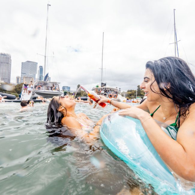 Two women enjoy a day in the water surrounded by boats. One woman pours a drink into the mouth of the other while people swim, and city buildings are visible in the background. It's a perfect scene for a Sydney boat party hire, showcasing fun and camaraderie under the sun.