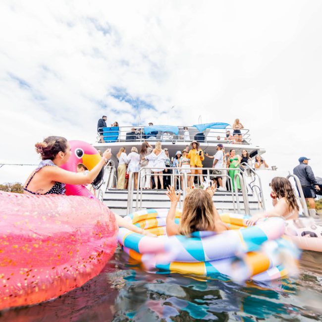 People are enjoying a Sydney boat party hire; some are on the boat while others float nearby in colorful inflatable rings.