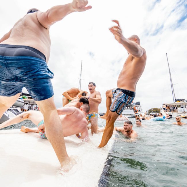 A group of men in swimwear engage in playful roughhousing on a floating platform surrounded by water, boats, and other people during a lively Sydney boat party hire.