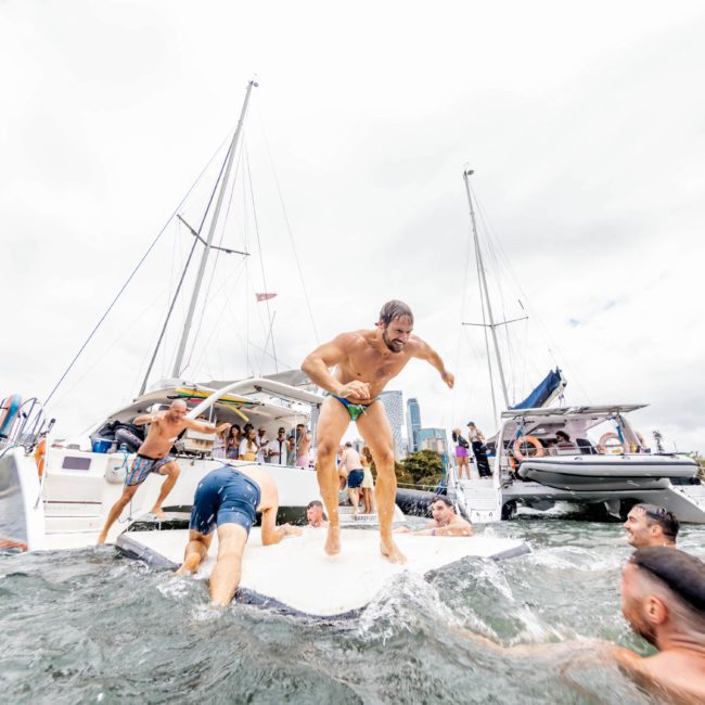 People are enjoying a swim and some are climbing back onto boats in a lively scene on the water, with multiple sailboats visible in the background. It's the perfect setting for a Sydney boat party hire or even a DJ boat hire Sydney experience.