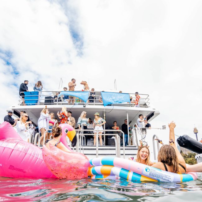 People on a boat and in the water enjoy a sunny day, with some on large inflatable pool floats, including a pink flamingo. The boat is lively and festive, reminiscent of a corporate boat event in Sydney.