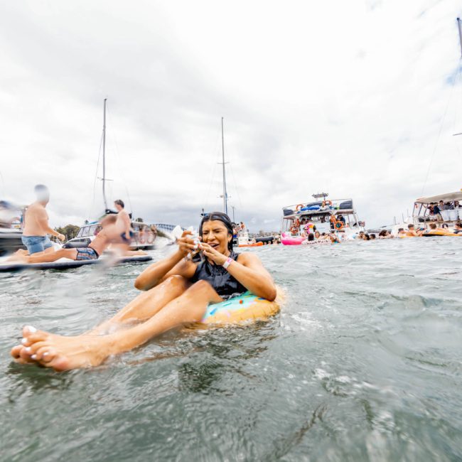 A person relaxes on an inflatable tube in the water, surrounded by boats and other people enjoying a vibrant Sydney boat party. The sky is cloudy.