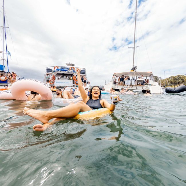 Several people are floating on inflatable items and swimming in the water near anchored boats under a cloudy sky. One person in the foreground holds up two drinks while floating on a yellow ring, enjoying what seems to be an incredible Sydney boat party hire experience.