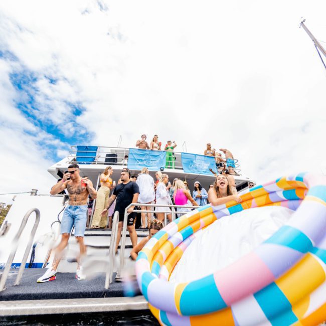 A group of people enjoying a catamaran party in Sydney. Some are dancing on the deck, while others are sliding down a floating water slide in the water. The sky is partly cloudy, setting a perfect scene for this corporate boat event.