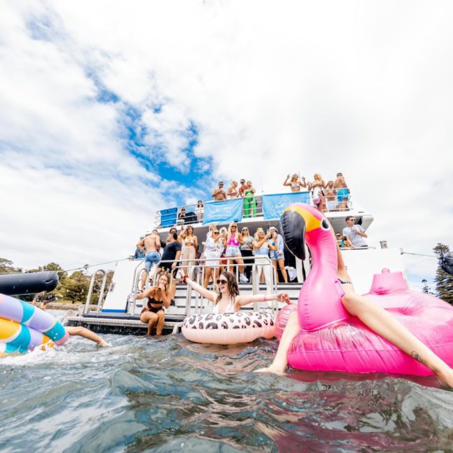 A group of people enjoy a corporate boat event in Sydney, with some on the deck and others in the water on inflatable floaties, including a large pink flamingo.