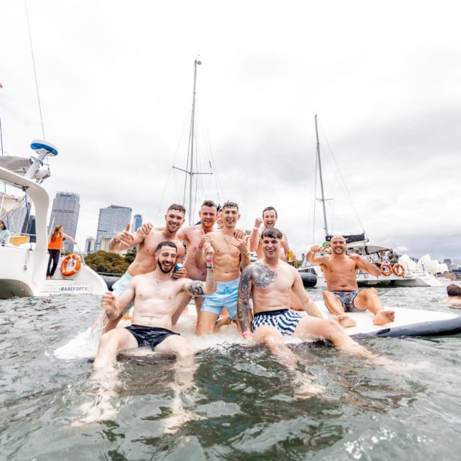 A group of men in swim attire enjoying time on an inflatable platform in the water, with sailboats and city buildings in the background on a cloudy day. Perfect for those looking to experience a Catamaran party Sydney offers.