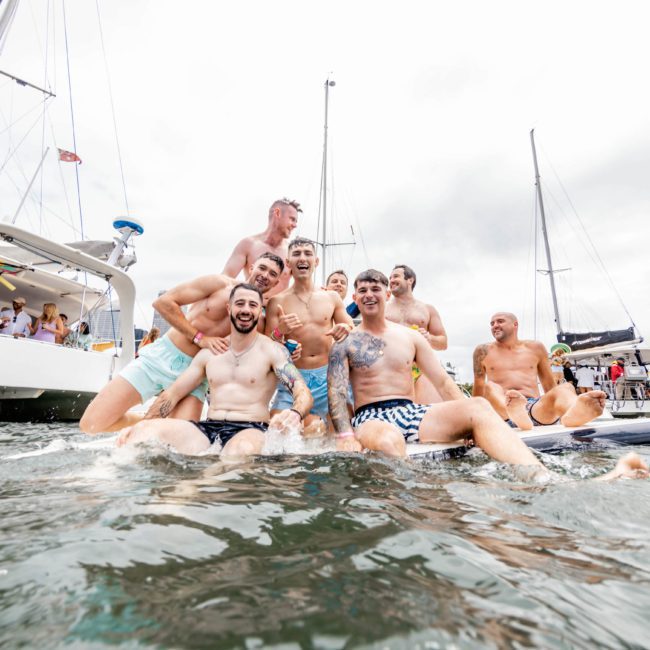 A group of people smiling and posing on a floating platform in the water near several sailboats on a cloudy day, enjoying a Sydney boat party hire.