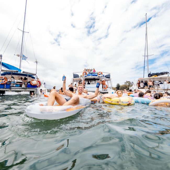 People relaxing on inflatable floats in the water, surrounded by boats at a Sydney boat party hire. They are enjoying beverages and the sunny weather.