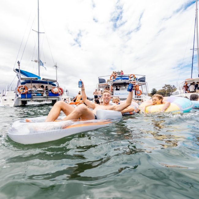 Two people smiling and holding drinks while lounging on an inflatable float, surrounded by several boats anchored in a waterway. Other people are visible on the boats and in the water, enjoying a vibrant Sydney boat party hire under the beautiful sun.