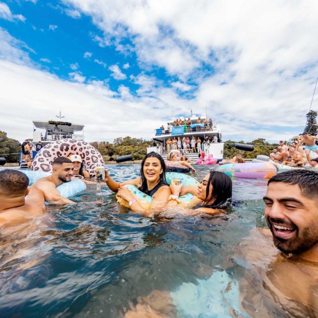 People enjoy a sunny day swimming and floating on inflatables in a body of water with boats anchored nearby and a group of people on a private yacht charter in the background.