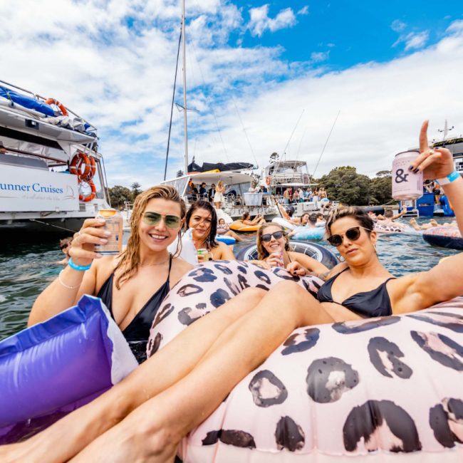 A group of people relax in inflatable pool floats in the water near boats, holding drinks and posing for the camera on a sunny day, enjoying a Catamaran party in Sydney.