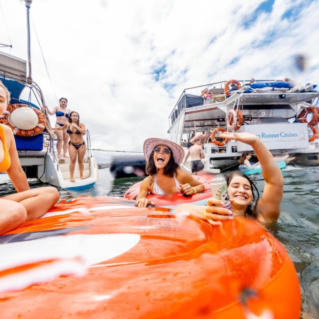 A group of people in swimwear enjoy a lively Catamaran party Sydney on the water near boats, with some on a large orange float and others on the deck. Some hold drinks, and one person is taking a photo.