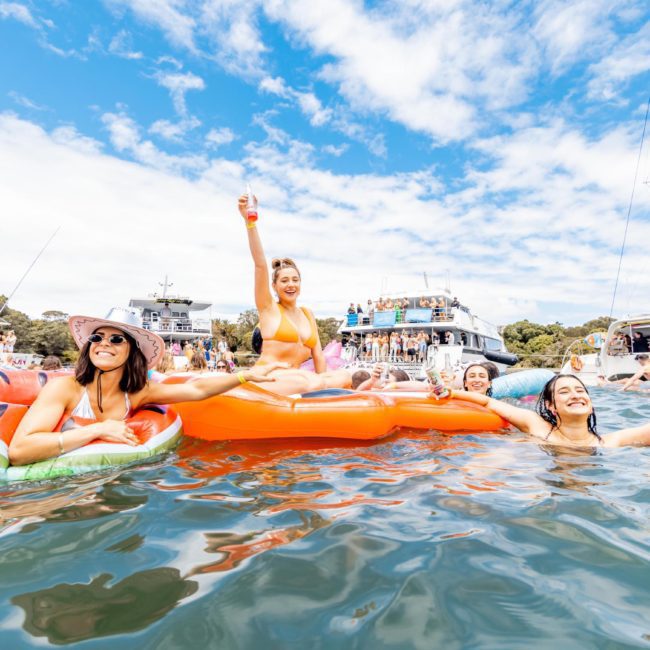 A group of people are enjoying a sunny day on the water with inflatables. One person is holding a drink aloft while others smile and relax on the luxury yacht hire Sydney. Boats and other partygoers are visible in the background.