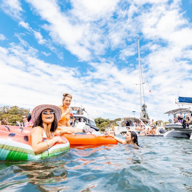 People enjoying a sunny day on the water with colorful floaties and boats around them. Some are swimming, while others are lounging and laughing, making it perfect for a catamaran party Sydney.