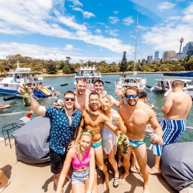 A group of people in swimsuits and casual clothing are smiling and holding drinks while standing on a boat with other boats and a city skyline in the background, enjoying a DJ boat hire Sydney.